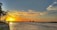 A scenic sunset over a calm body of water with a vibrant orange and yellow sky. Chinese fishing nets are visible on the left, along with a person standing on the shore. Distant buildings and silhouettes of trees line the horizon.