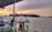 A large cargo ship is moving across a body of water, loaded with multicolored shipping containers. In the foreground, a wooden dock and some vibrant orange and green plants add a splash of color. The sky is overcast with a mix of gray clouds and a golden sunset.