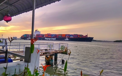 A large cargo ship is moving across a body of water, loaded with multicolored shipping containers. In the foreground, a wooden dock and some vibrant orange and green plants add a splash of color. The sky is overcast with a mix of gray clouds and a golden sunset.