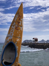 A worn-out yellow surfboard with the words 'Stand Up Kite' is positioned on the left side against a backdrop of a rocky pier. In the distance, there is a small lifeguard tower and rugged hills beyond. The ocean waves crash gently against the pier, under a sky partly covered with scattered white clouds.