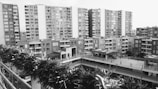 A residential area features several mid-rise apartment buildings with multiple balconies. The view is slightly elevated, overlooking a green fence in the foreground, which appears to be part of a terrace or balcony. The scene lacks any noticeable human activity, conveying quietness.