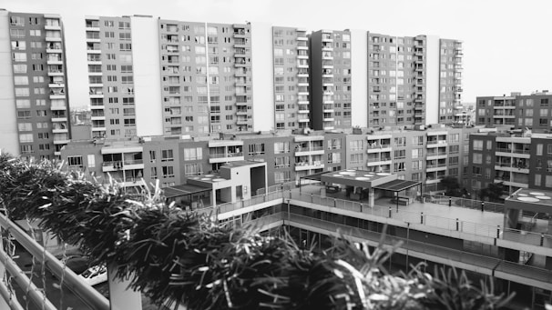 A residential area features several mid-rise apartment buildings with multiple balconies. The view is slightly elevated, overlooking a green fence in the foreground, which appears to be part of a terrace or balcony. The scene lacks any noticeable human activity, conveying quietness.