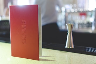 A red menu labeled 'DRINKS' stands upright on a bar counter. Next to it, a metal jigger is placed, likely used for measuring cocktails or beverages. The background includes a blurred setting with bar equipment and possible patrons.