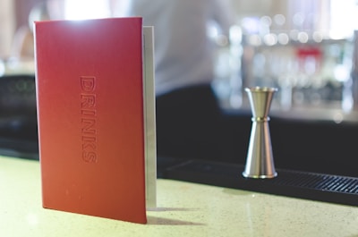 A red menu labeled 'DRINKS' stands upright on a bar counter. Next to it, a metal jigger is placed, likely used for measuring cocktails or beverages. The background includes a blurred setting with bar equipment and possible patrons.