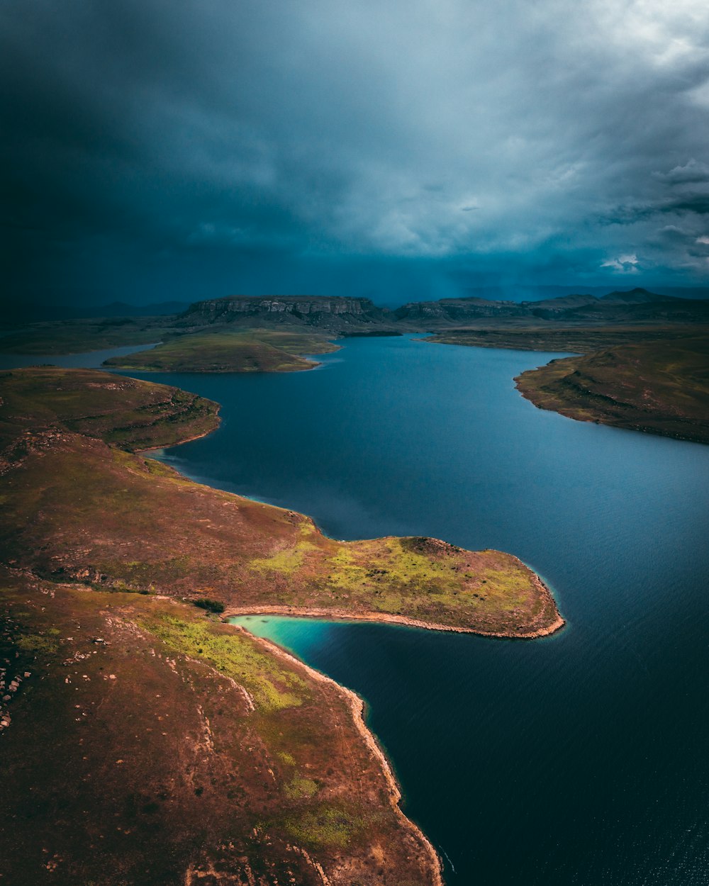 Fotografía aérea de un cuerpo de agua con un campo marrón y verde bajo nubes dramáticas