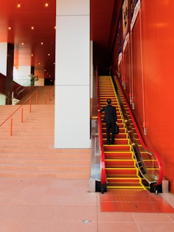 A person dressed in formal attire ascends an escalator inside a modern building. The interior features bold red and white colors with red railings along a staircase parallel to the escalator. Overhead lighting provides a warm ambiance, and advertisements are visible on the wall next to the escalator.