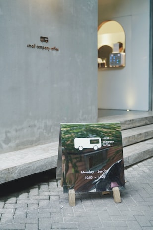 A small coffee shop entrance with a minimalistic sign on the wall reading 'small company coffee.' In front of it, a reflective sandwich board displays the same logo along with opening hours, Monday to Sunday from 10:00 to 19:00. The sidewalk is paved with bricks, leading to a modern interior visible through the door.