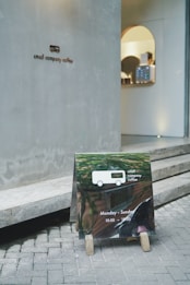 A small coffee shop entrance with a minimalistic sign on the wall reading 'small company coffee.' In front of it, a reflective sandwich board displays the same logo along with opening hours, Monday to Sunday from 10:00 to 19:00. The sidewalk is paved with bricks, leading to a modern interior visible through the door.