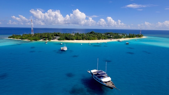 A tranquil, tropical island surrounded by clear blue waters. Several boats are anchored near the shoreline, and lush greenery covers much of the island. Communication towers are visible on either side of the island. The sky is clear with scattered clouds, adding to the serene ambiance.