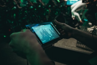 Close-up view of a person holding a tablet-like device with a screen displaying a group of people. In the background, there are several stationary bikes, and a person wearing white leggings is partially visible. The lighting is dim, with a greenish hue dominating the scene.