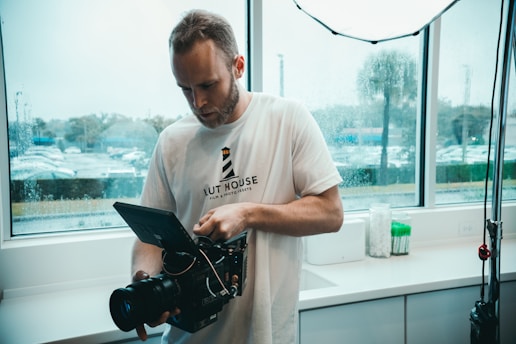 A man in a white t-shirt is holding a professional video camera, standing indoors by a window with a rainy view outside. The room appears to be well-lit with fluorescent lighting.