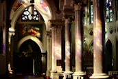 Intricately designed columns and arches with colorful stained glass windows cast vibrant reflections inside a church. Illuminated religious artwork and iconography adorn the walls, creating a serene and spiritual atmosphere.