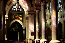Intricately designed columns and arches with colorful stained glass windows cast vibrant reflections inside a church. Illuminated religious artwork and iconography adorn the walls, creating a serene and spiritual atmosphere.