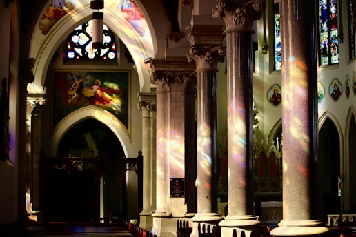 Intricately designed columns and arches with colorful stained glass windows cast vibrant reflections inside a church. Illuminated religious artwork and iconography adorn the walls, creating a serene and spiritual atmosphere.