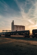 A large industrial building labeled 'Port of Sacramento' stands prominently in the background. In the foreground, a long black train with tanker cars stretches across the scene. The sky has a mix of blue and orange hues, indicating either sunrise or sunset, adding a warm glow to the image.