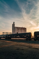 A large industrial building labeled 'Port of Sacramento' stands prominently in the background. In the foreground, a long black train with tanker cars stretches across the scene. The sky has a mix of blue and orange hues, indicating either sunrise or sunset, adding a warm glow to the image.