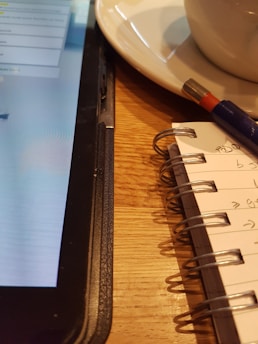 A close-up view of a tablet, a notebook, and a pen on a wooden table. The tablet displays text on the screen, partially visible. The notebook is spiral-bound with handwritten notes, and a pen rests on top.
