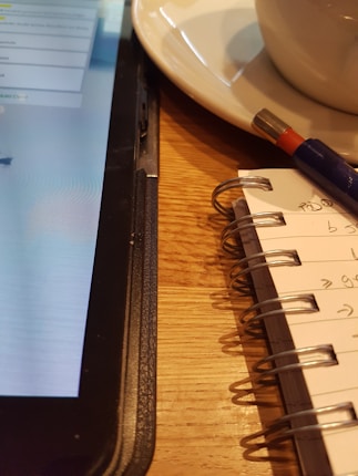 A close-up view of a tablet, a notebook, and a pen on a wooden table. The tablet displays text on the screen, partially visible. The notebook is spiral-bound with handwritten notes, and a pen rests on top.