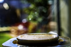 A shallow depth of field captures a close-up view of a steaming beverage or dish in a shiny, metallic container. The background is blurred, featuring soft bokeh and greenery, which suggests an outdoor setting.