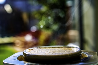 A shallow depth of field captures a close-up view of a steaming beverage or dish in a shiny, metallic container. The background is blurred, featuring soft bokeh and greenery, which suggests an outdoor setting.