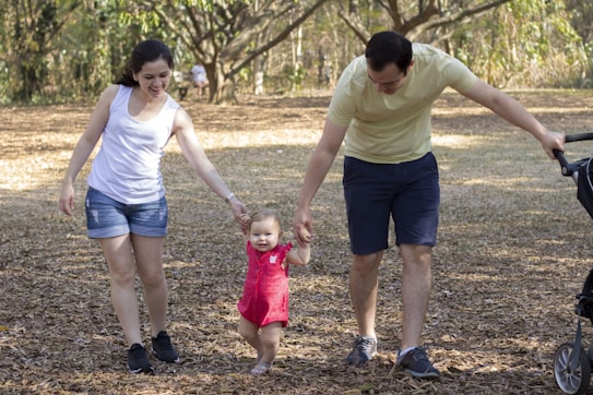 A toddler, wearing a bright pink outfit, is joyfully walking with assistance from two adults holding their hands. The setting is an outdoor park with scattered fallen leaves and trees in the background. The adults appear to be smiling, and a stroller is visible on the side.