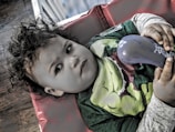 A child with curly hair is lying on a red cushion, holding a blue object in their hands. The child is wearing a green shirt with a superhero design and appears to be gazing thoughtfully.