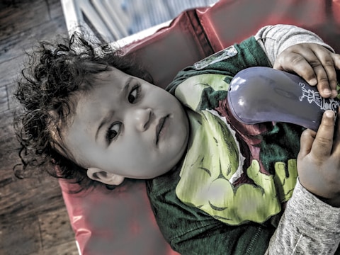 A child with curly hair is lying on a red cushion, holding a blue object in their hands. The child is wearing a green shirt with a superhero design and appears to be gazing thoughtfully.