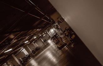 A dimly lit underground parking garage with a reflective floor. The space is lined with parked motorcycles and scooters on the sides. Structural beams are visible on the ceiling, and there are columns dividing the space with alphanumeric designations like 'A9' marked on them.
