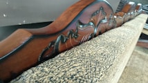 Ornately carved wooden headboard with a smooth finish, featuring intricate floral designs. It rests against a textured, patterned fabric, likely part of a piece of furniture such as a sofa or a bed.