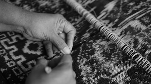 Hands carefully engage in fabric weaving, manipulating threads with precision on a loom. The monochromatic pattern on the textile reveals intricate geometric designs, reflecting traditional craftsmanship.