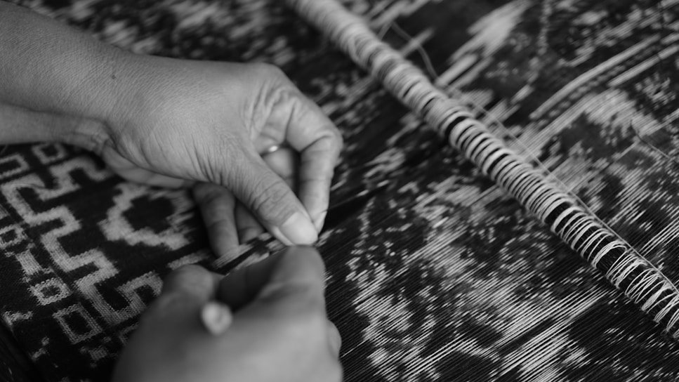Hands carefully engage in fabric weaving, manipulating threads with precision on a loom. The monochromatic pattern on the textile reveals intricate geometric designs, reflecting traditional craftsmanship.