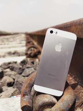 A smartphone is positioned upright on a metal railroad track, surrounded by stones. The phone's back displays the Apple logo and 'iPhone' text. The scene suggests an outdoor setting, possibly along a railway line.