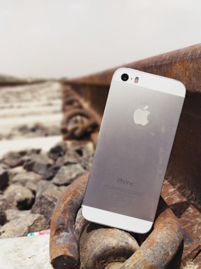 A smartphone is positioned upright on a metal railroad track, surrounded by stones. The phone's back displays the Apple logo and 'iPhone' text. The scene suggests an outdoor setting, possibly along a railway line.