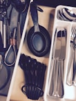 A neatly organized kitchen drawer containing various utensils arranged in separate compartments. Measuring spoons and cups in black plastic are placed alongside metal scissors and other kitchen tools. A tray with stainless steel cutlery including knives, forks, and spoons is visible.