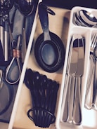 A neatly organized kitchen drawer containing various utensils arranged in separate compartments. Measuring spoons and cups in black plastic are placed alongside metal scissors and other kitchen tools. A tray with stainless steel cutlery including knives, forks, and spoons is visible.