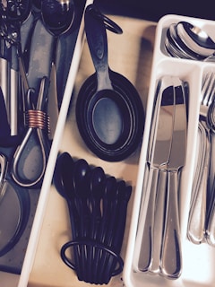 A neatly organized kitchen drawer containing various utensils arranged in separate compartments. Measuring spoons and cups in black plastic are placed alongside metal scissors and other kitchen tools. A tray with stainless steel cutlery including knives, forks, and spoons is visible.