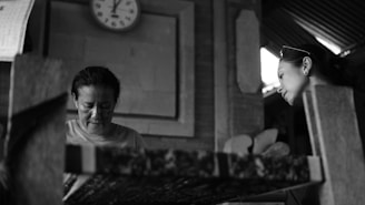 Two individuals are engaged in a concentrated activity in an interior setting. A woman is positioned to the left of the image, focused on her work, while another woman is on the right, observing with interest. A clock displaying a time of 3:10 is mounted on the wall behind them.