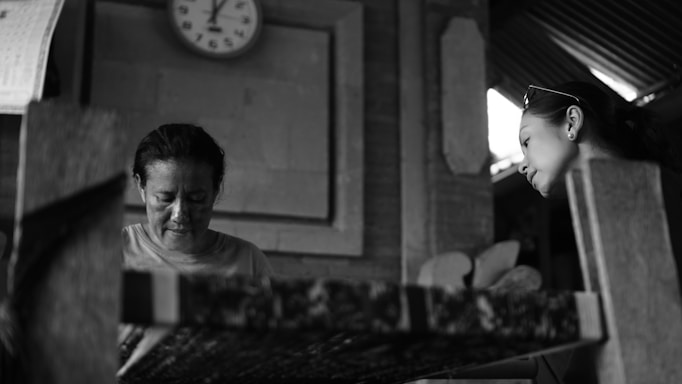 Two individuals are engaged in a concentrated activity in an interior setting. A woman is positioned to the left of the image, focused on her work, while another woman is on the right, observing with interest. A clock displaying a time of 3:10 is mounted on the wall behind them.