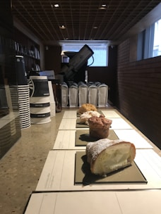 A counter displays several pastry items arranged in a line, each on a rectangular black tray, with a creamy filling visible in one pastry. In the background, there are stacks of paper coffee cups and some appliances, possibly a coffee machine. The setting appears to be well-lit and features a structured ceiling and brick walls.