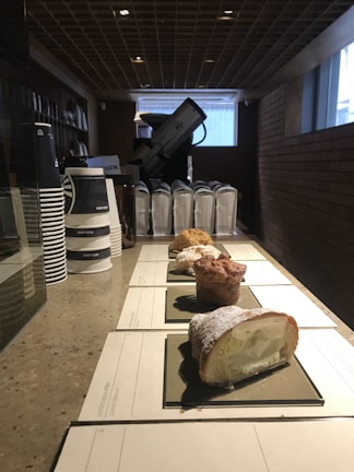 A counter displays several pastry items arranged in a line, each on a rectangular black tray, with a creamy filling visible in one pastry. In the background, there are stacks of paper coffee cups and some appliances, possibly a coffee machine. The setting appears to be well-lit and features a structured ceiling and brick walls.