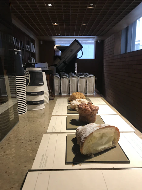 A counter displays several pastry items arranged in a line, each on a rectangular black tray, with a creamy filling visible in one pastry. In the background, there are stacks of paper coffee cups and some appliances, possibly a coffee machine. The setting appears to be well-lit and features a structured ceiling and brick walls.