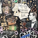 A collage featuring various electronic components, wires, and circuit boards arranged artistically on a dark background. A vintage photograph of a man wearing a tuxedo is incorporated into the scene, positioned to the right, with a cardboard crown on his head. The composition includes stenciled graphics, stars, hearts, and graffiti art, with text that reads 'Welcome to the Technological Babylonian Era'.