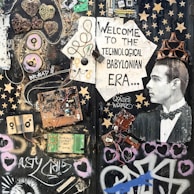 A collage featuring various electronic components, wires, and circuit boards arranged artistically on a dark background. A vintage photograph of a man wearing a tuxedo is incorporated into the scene, positioned to the right, with a cardboard crown on his head. The composition includes stenciled graphics, stars, hearts, and graffiti art, with text that reads 'Welcome to the Technological Babylonian Era'.