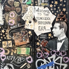 A collage featuring various electronic components, wires, and circuit boards arranged artistically on a dark background. A vintage photograph of a man wearing a tuxedo is incorporated into the scene, positioned to the right, with a cardboard crown on his head. The composition includes stenciled graphics, stars, hearts, and graffiti art, with text that reads 'Welcome to the Technological Babylonian Era'.