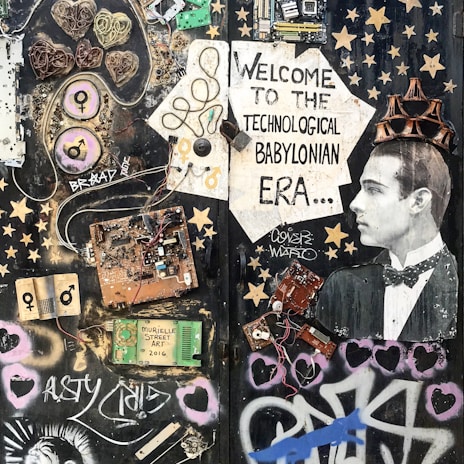 A collage featuring various electronic components, wires, and circuit boards arranged artistically on a dark background. A vintage photograph of a man wearing a tuxedo is incorporated into the scene, positioned to the right, with a cardboard crown on his head. The composition includes stenciled graphics, stars, hearts, and graffiti art, with text that reads 'Welcome to the Technological Babylonian Era'.