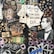 A collage featuring various electronic components, wires, and circuit boards arranged artistically on a dark background. A vintage photograph of a man wearing a tuxedo is incorporated into the scene, positioned to the right, with a cardboard crown on his head. The composition includes stenciled graphics, stars, hearts, and graffiti art, with text that reads 'Welcome to the Technological Babylonian Era'.