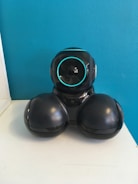 A black robotic device with a spherical head featuring a glowing blue ring. The body is rounded and appears to rest on two broad, bulbous sections. The device is positioned against a bright blue wall and placed on a white surface.