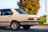 A beige vintage sedan is parked on a paved surface with a lush green landscape and a tree in the background. The car features a distinctive chrome trim, classic styling, and European license plate.