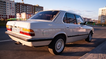 A classic white sedan is parked on a city street, reflecting the late afternoon sunlight. The background features several mid-rise apartment buildings in a modern urban setting. The car is well-maintained, showcasing its sleek lines and polished surface.