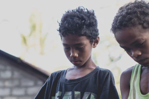 Two young boys are standing close together with their heads bowed. The background appears to be slightly blurred, with elements of a building partially visible. The boys have curly hair and are wearing casual clothing. The lighting is natural, suggesting an outdoor setting.
